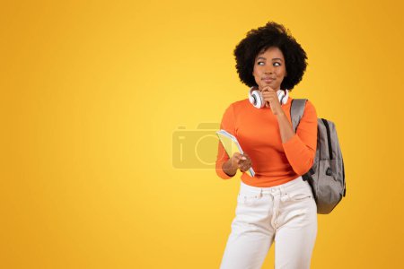 Téléchargez les photos : Étudiant afro-américain réfléchi avec des cheveux bouclés et des écouteurs autour du cou, tenant un cahier et un stylo, portant un haut orange et un pantalon blanc avec un sac à dos, sur un fond jaune - en image libre de droit