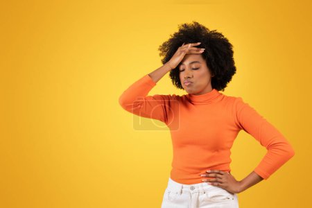 Foto de Una joven afroamericana con expresión reflexiva se levanta sobre un fondo amarillo, tocándose la frente con la mano, vistiendo un cuello alto naranja y pantalones blancos - Imagen libre de derechos