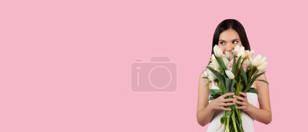 Téléchargez les photos : Radieuse jeune femme partiellement cachée derrière un bouquet de tulipes fraîches, regardant de côté l'espace libre, exprimant joie et ludique, isolée sur fond rose doux pour la Journée de la femme - en image libre de droit