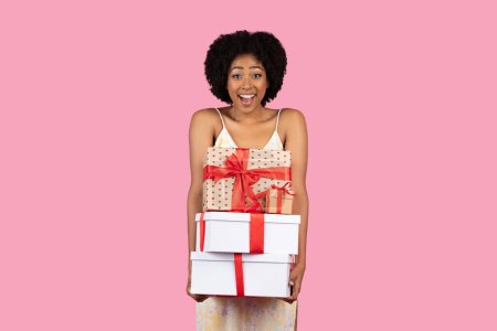 Foto de Mujer afroamericana milenaria llena de alegría con el pelo rizado, sosteniendo una pila de cajas de regalo de varios tamaños envueltas con cintas rojas, expresando emoción sobre un fondo rosa - Imagen libre de derechos
