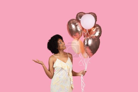 Téléchargez les photos : Femme afro-américaine joyeuse millénaire aux cheveux bouclés, regardant vers le haut des ballons en or rose et blanc dans sa main, avec un sourire doux, debout sur un fond rose - en image libre de droit