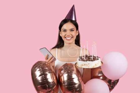 Strahlende junge Frau mit Partyhut hält Geburtstagstorte mit brennenden Kerzen und glänzenden Luftballons in der Hand und posiert fröhlich mit ihrem Smartphone auf rosa Hintergrund