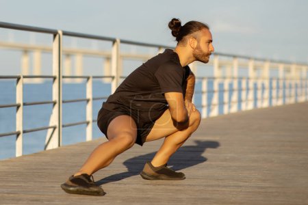 Photo for Young man in black fitwear engages in outdoor morning workout by the pier, doing deep lunge exercise that promotes muscle engagement and overall fitness for healthy lifestyle, full length - Royalty Free Image