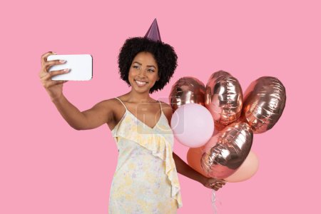 Vibrant millennial African American woman with curly hair and a party hat taking a selfie, with a bright smile, holding rose gold balloons on a pink celebration background