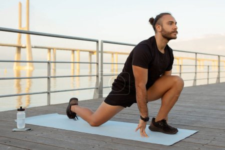 Photo for Outdoor fitness. Motivated young man in sporty attire doing leg stretches on mat outside. Guy making kneeling hip flexor exercise during stretching training on pier. Workout, wellbeing - Royalty Free Image