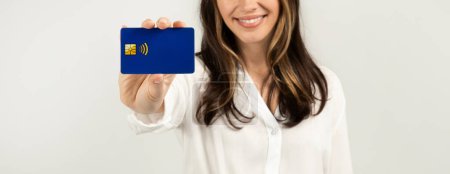Photo for Cheerful european woman in a white blouse holding a blue credit card towards the camera, with a focus on the card, symbolizing financial transactions and consumerism, studio - Royalty Free Image