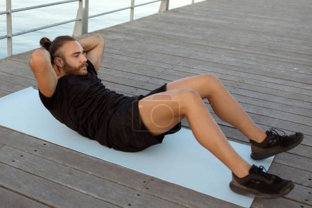 Seaside workout session. Active sporty man in sportswear lies on a mat and doing abs crunches, engaging in abdominal exercises during training outside. Strong body, fitness motivation
