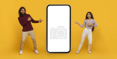 Téléchargez les photos : Belle offre en ligne. Cool élégant jeune Indien homme et femme pointant vers grand smartphone avec blanc écran blanc maquette copie espace entre eux, fond jaune, bannière web - en image libre de droit