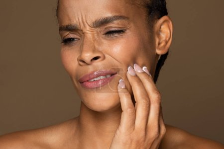 Photo for Closeup of african american distressed woman pressing her cheek with pained expression, suffering toothache, facial discomfort, brown studio background - Royalty Free Image