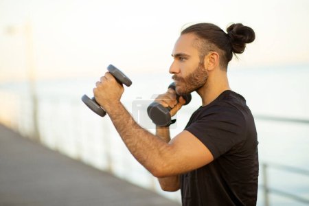 Photo for Side view portrait of determined European sporty guy in sportswear, boxing outdoors by the seaside, having bodybuilding workout and strengthening his arms. Strength and active lifestyle - Royalty Free Image