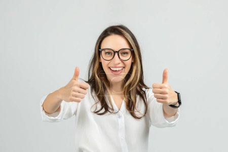Photo for A smiling european young woman with long hair and glasses gives two enthusiastic thumbs up, wearing a white blouse and smartwatch, showing approval or success, studio, close up - Royalty Free Image