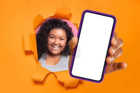 Foto de Alegre afroamericana más tamaño joven con el pelo rizado mirando a través de un papel naranja desgarrado, presentando un teléfono inteligente con una pantalla en blanco, perfecto para diseños de maquetas - Imagen libre de derechos