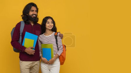 Téléchargez les photos : Curieux jeune Indien homme et femme avec des sacs à dos derrière le dos et des livres dans leurs mains étudiants étreignant et regardant l'espace de copie sur fond jaune, bannière web pour l'éducation internationale - en image libre de droit