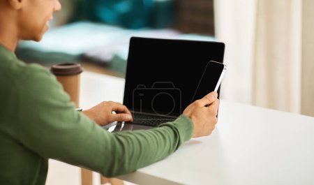 Photo for Over shoulder view black guy independent contractor using laptop computer and smartphone with blank screen, sitting at cafe, drinking coffee. Technologies and business concept - Royalty Free Image