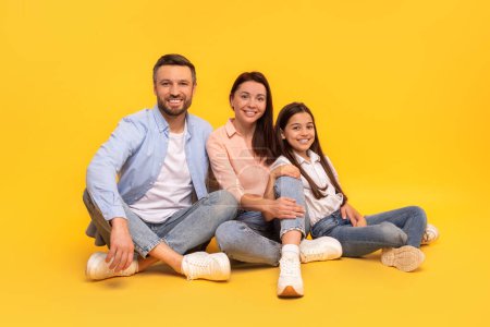 Téléchargez les photos : Heureuse famille de trois personnes vêtues de façon décontractée partageant un moment de convivialité, assises dans une pose détendue sur fond de studio jaune, parents et fillette souriant à la caméra - en image libre de droit