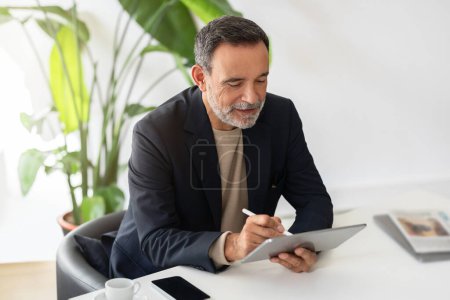 Foto de Hombre de negocios caucásico maduro enfocado en un traje inteligente que trabaja intensamente en una tableta con un lápiz, con una taza de café y un teléfono inteligente en la mesa, en una oficina luminosa con vegetación - Imagen libre de derechos