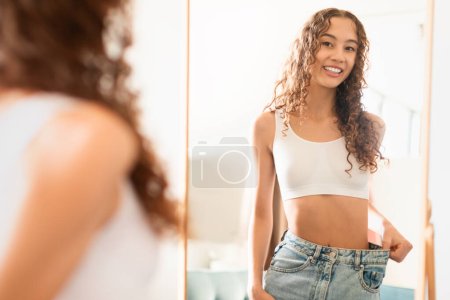 Téléchargez les photos : Adolescente caucasienne regardant miroir à la maison, portant un jean de grande taille, mettant joyeusement en valeur le succès de la perte de poids. Dame heureuse à la taille mince sourit à la réflexion, la promotion d'un mode de vie sain - en image libre de droit