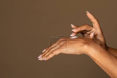 Photo for Graceful black female hands with manicured nails applying dab of moisturizing cream on the back of hand against warm, neutral brown background - Royalty Free Image