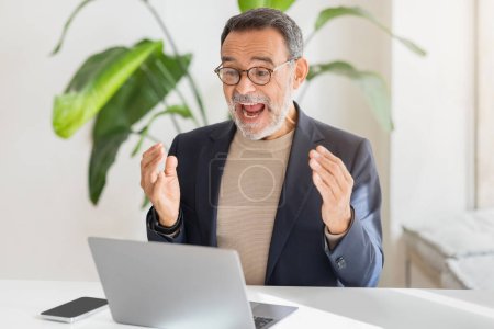 Photo for An exuberant happy caucasian middle-aged businessman with glasses joyfully reacts during a video call on his laptop in a bright, plant-adorned office space. Win emotions, success, good news - Royalty Free Image