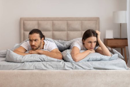 Couple lies in bed with troubled expressions, facing away from each other, suggesting disagreement or emotional distance in their relationship