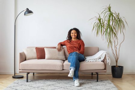 Foto de Joven y alegre mujer negra sentada cómodamente en un sofá moderno con cojines, feliz mujer afroamericana milenaria posando en una sala de estar bien iluminada, sonriendo a la cámara, espacio para copiar - Imagen libre de derechos