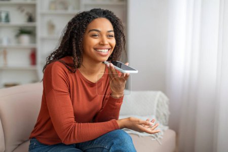 Téléchargez les photos : Technologies modernes. Portrait d'une jeune femme noire heureuse enregistrant un message vocal sur un téléphone intelligent tout en se relaxant à la maison, souriante afro-américaine naviguant sur assistant virtuel sur téléphone mobile - en image libre de droit