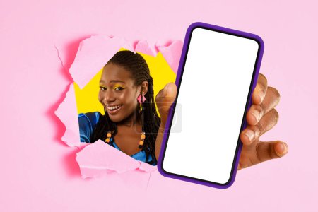Foto de Mujer afroamericana adulta alegre con cabello trenzado y pendientes coloridos que dan un pulgar hacia arriba a través de un papel roto rosa, mostrando un teléfono inteligente con una pantalla en blanco para mostrar - Imagen libre de derechos