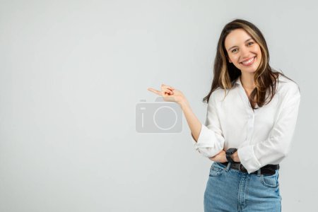 Foto de Una simpática joven europea sonriente con una sonrisa radiante apunta hacia un lado mientras usa una blusa blanca crujiente y vaqueros azules, sugiriendo una opción o dirección, estudio - Imagen libre de derechos
