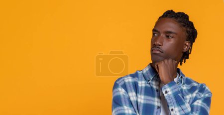 Foto de Pensativo chico afroamericano mirando a un lado en el espacio de copia y tocando la barbilla, reflexivo joven negro pensando en la oferta interesante, de pie aislado en el fondo del estudio amarillo, Panorama - Imagen libre de derechos