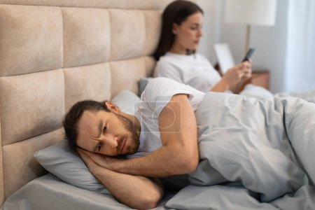 Foto de El hombre parece preocupado e incapaz de dormir mientras yace en la cama, mientras que la mujer, aparentemente indiferente usando su teléfono, retrata el tema moderno de la interferencia de la tecnología en la intimidad - Imagen libre de derechos