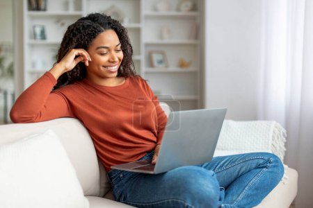Foto de Mujer negra sonriente que se relaja con el ordenador portátil en el sofá en casa, Mujer afroamericana joven y feliz sentada en el sofá en la sala de estar, viendo películas o navegando por Internet en la computadora, Copiar el espacio - Imagen libre de derechos