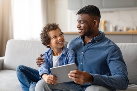 Téléchargez les photos : Le père afro-américain et son fils passent un bon moment ensemble, partageant une tablette numérique et échangeant des sourires sur un canapé confortable dans un salon lumineux - en image libre de droit