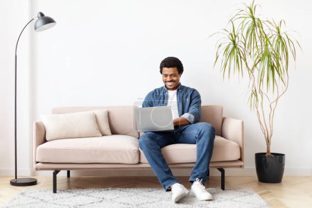 Foto de Concepto Independiente. Guapo joven negro hombre usando el ordenador portátil mientras está sentado en el sofá en casa, Millennial chico afroamericano trabajando de forma remota en la computadora en la sala de estar, Disfrutando del trabajo en línea, Copiar el espacio - Imagen libre de derechos