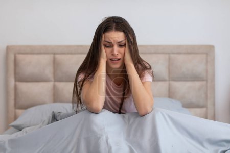 Photo for Young woman sits upright in bed, her hands clasping her head, her expression one of pain or deep concern, evoking sense of acute headache, stress, or emotional distress - Royalty Free Image