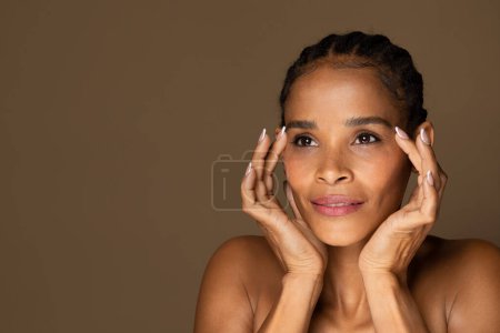 Photo for Radiant black middle aged woman gently framing her face with her hands, showcasing moment of beauty and contemplation against brown background, free space - Royalty Free Image