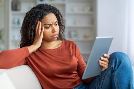 Photo for Concerned young black woman looking at digital tablet and touching head in distress, upset african american female reading bad news online, sitting on couch in living room at home, closeup - Royalty Free Image