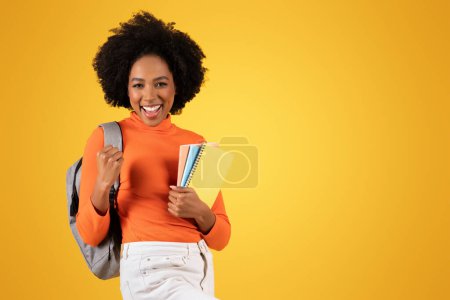 Téléchargez les photos : Jeune étudiant afro-américain excité avec un sourire large, tenant des carnets et portant un col roulé orange avec un sac à dos gris, sur un fond jaune joyeux - en image libre de droit