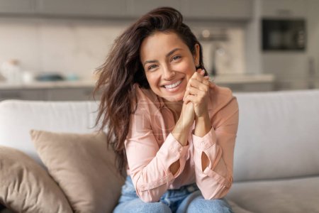 Photo for Radiant woman with tousled hair, wearing pink blouse, resting on comfy sofa with joyful smile and clasped hands, exuding happiness and enjoying time at home - Royalty Free Image