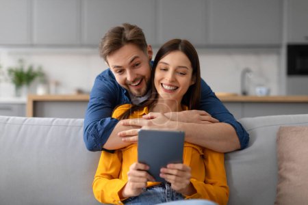 Fröhliche Ehepartner mit digitalem Tablet beschäftigt, Mann umarmt Frau herzlich von hinten, beide zeigen helles Lächeln, Videotelefonie mit Familie oder Surfen im Internet
