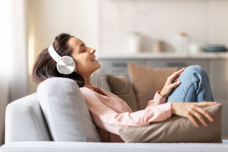 Téléchargez les photos : Contente jeune femme allongée confortablement sur le canapé, les yeux fermés, perdue dans la musique grâce à ses écouteurs blancs élégants, se sentant sereine, vue de côté - en image libre de droit