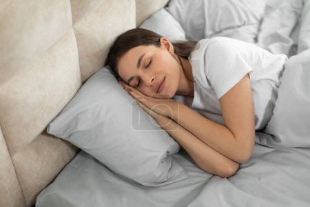 Ruhige junge Frau liegt bequem im Bett, ihr Gesichtsausdruck ist ruhig, während sie einen tiefen, erholsamen Schlaf genießt, der Ruhe und Entspannung suggeriert, Schlafzimmereinrichtung