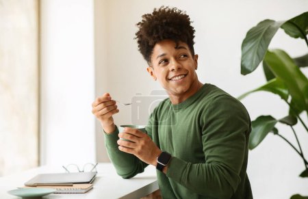 Photo for Stylish african american guy with curly hair drinking cappuccino while working at cafe, sitting at table next to window at coffee shop, holding mug and spoon, looking at copy space - Royalty Free Image
