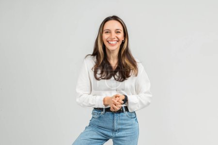 Photo for A cheerful european young woman with long hair wearing a white blouse and blue jeans stands with clasped hands against a plain background, exuding a friendly demeanor, studio - Royalty Free Image
