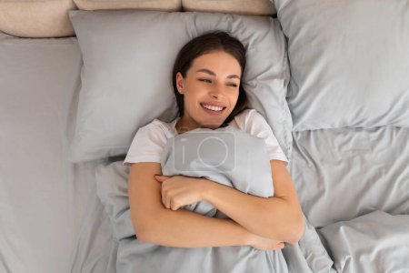 Contented young woman lies in bed, clutching pillow with joyful smile, exuding sense of happiness and comfort in her serene bedroom environment