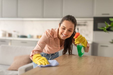 Foto de Mujer joven alegre en ropa casual y guantes amarillos pulveriza felizmente limpiador en la mesa de la cocina de madera, dedicándose a las tareas domésticas - Imagen libre de derechos