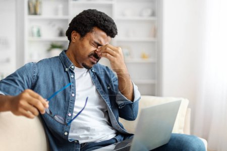Téléchargez les photos : Homme noir fatigué frottant les yeux, montrant des signes de fatigue ou de stress après avoir utilisé un ordinateur portable à la maison, homme afro-américain épuisé assis sur le canapé dans le salon, ayant la fatigue oculaire, copier l'espace - en image libre de droit