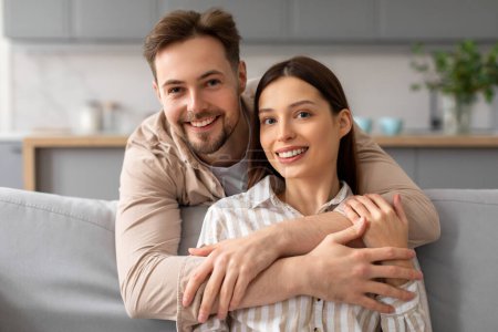 Photo for Smiling young man hugging his girlfriend from behind on couch, showing moment of affection and closeness in bright, cozy living room, couple looking at camera - Royalty Free Image