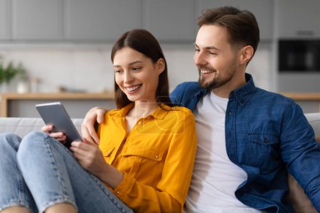 Fröhliche junge Frau und Mann genießen das gemeinsame Surfen auf dem digitalen Tablet, Online-Shopping oder Videotelefonie, bequem auf der Couch im Wohnzimmer sitzend