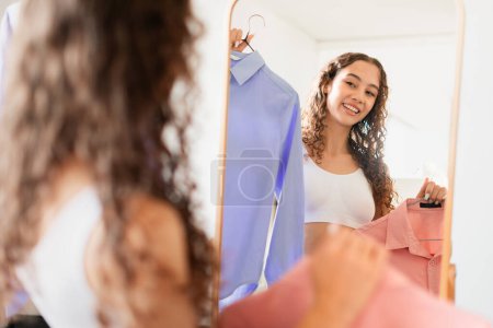 Caucásico adolescente chica felizmente elegir camisa de moda sosteniendo prendas y mirando el espejo en casa. Joven comprador alegre mostrando concepto de elección personal en prendas de vestir. Moda y estilo individual