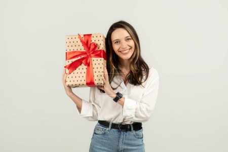 Téléchargez les photos : Charmante femme européenne souriante avec un sourire rayonnant tenant un cadeau magnifiquement enveloppé avec un arc rouge, suggérant une occasion festive ou un geste réfléchi de don, studio - en image libre de droit